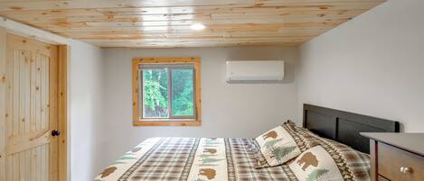 A bedroom with a wooden ceiling, a window with a view of trees, a bed with a patterned bedspread, and a bedside table with a remote control.
