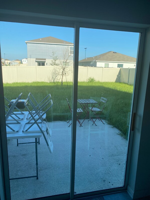 A view through a glass door of a backyard with a patio table and chairs, a grassy area, and a building in the background.