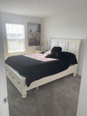 A bedroom with a bed, a nightstand, a window with blinds, a wall-mounted painting, and a door.
