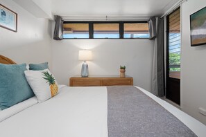 A bedroom with a bed, pillows, a bedside table with a lamp, a small plant, and a window with curtains. - 1BD Condo with Ocean & Sunset Views Near Beach -Kahana Villas F407 (Lahaina)