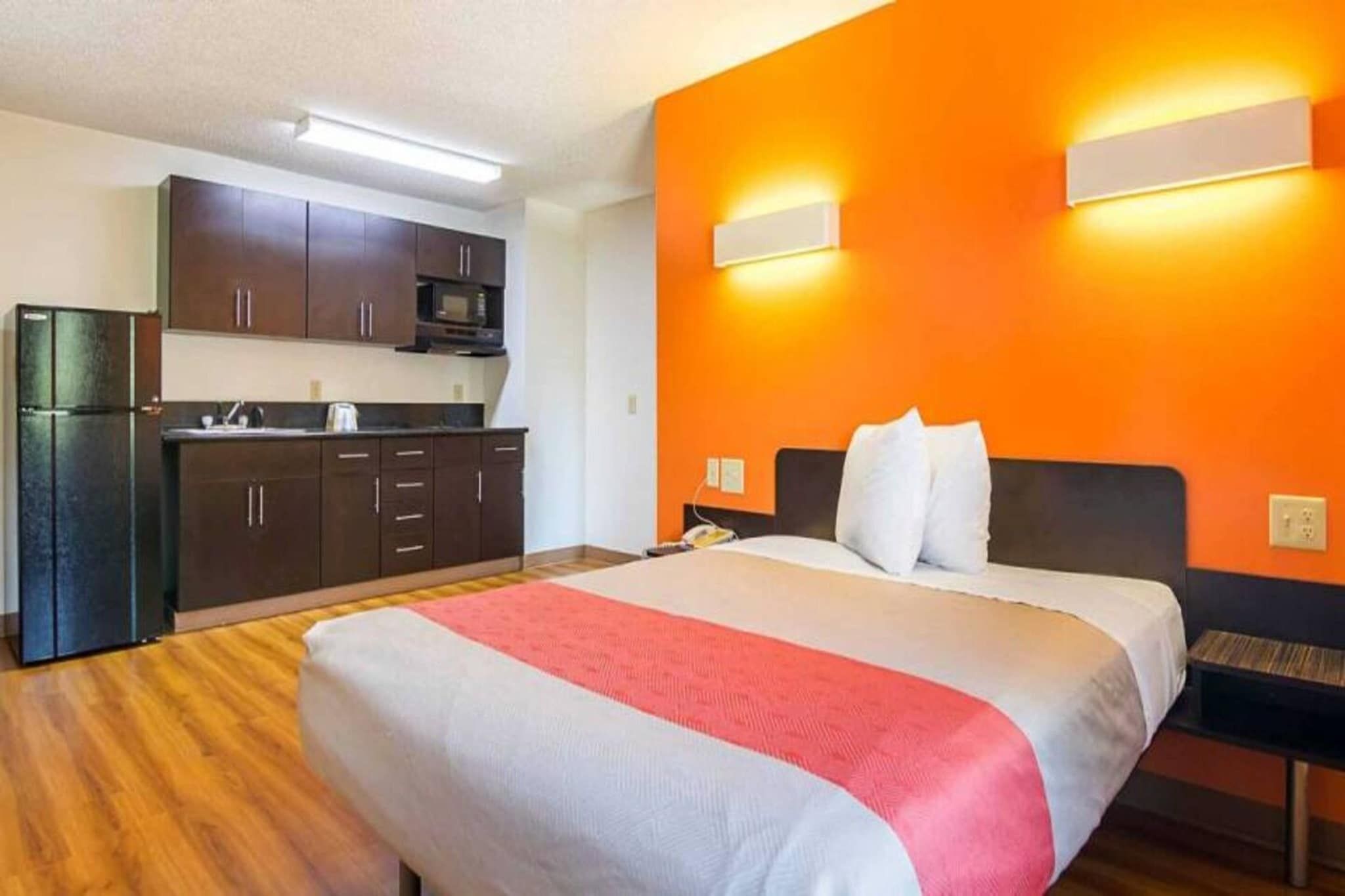 A hotel room with a bed, a nightstand, a desk, and a kitchenette.