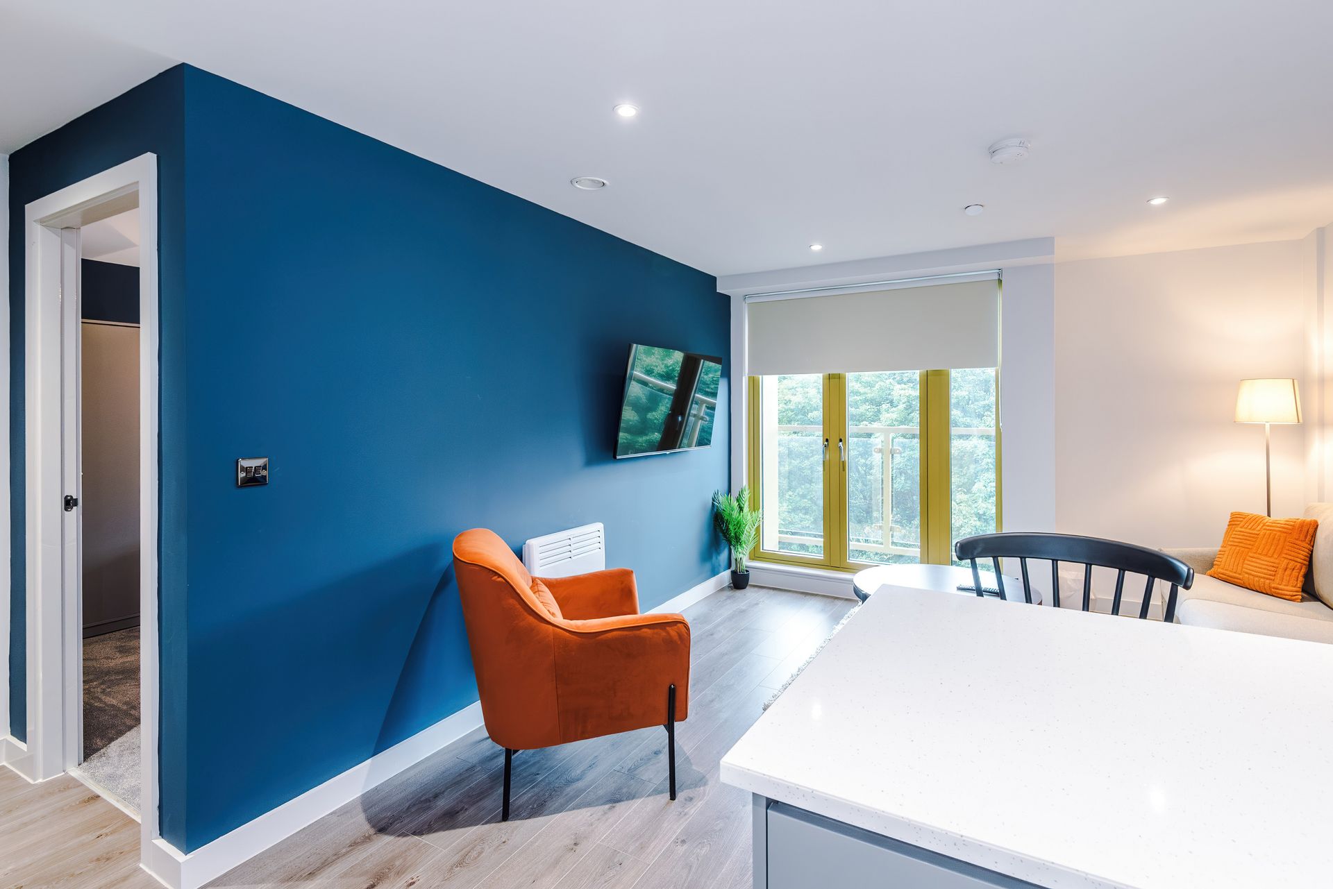A modern living room with a blue accent wall, a flat-screen TV, a dining area with a white table, and a view through a large window.