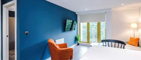 A modern living room with a blue accent wall, a flat-screen TV, a dining area with a white table, and a view through a large window.