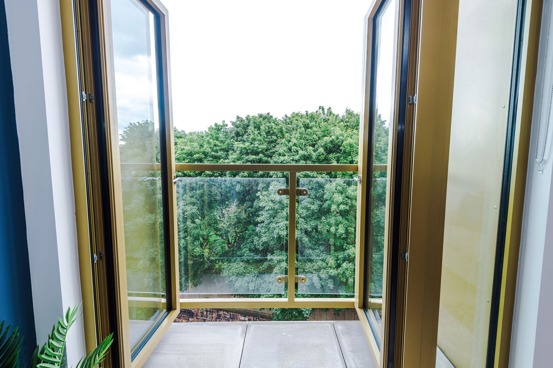 Un balcon doté d’une balustrade en verre, offrant une vue sur une zone boisée, et une porte donnant sur l’intérieur.
