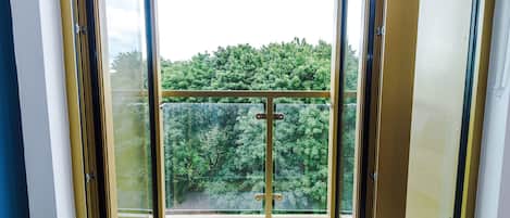 Un balcón con barandilla de vidrio, vista a zona boscosa y puerta que da al interior.
