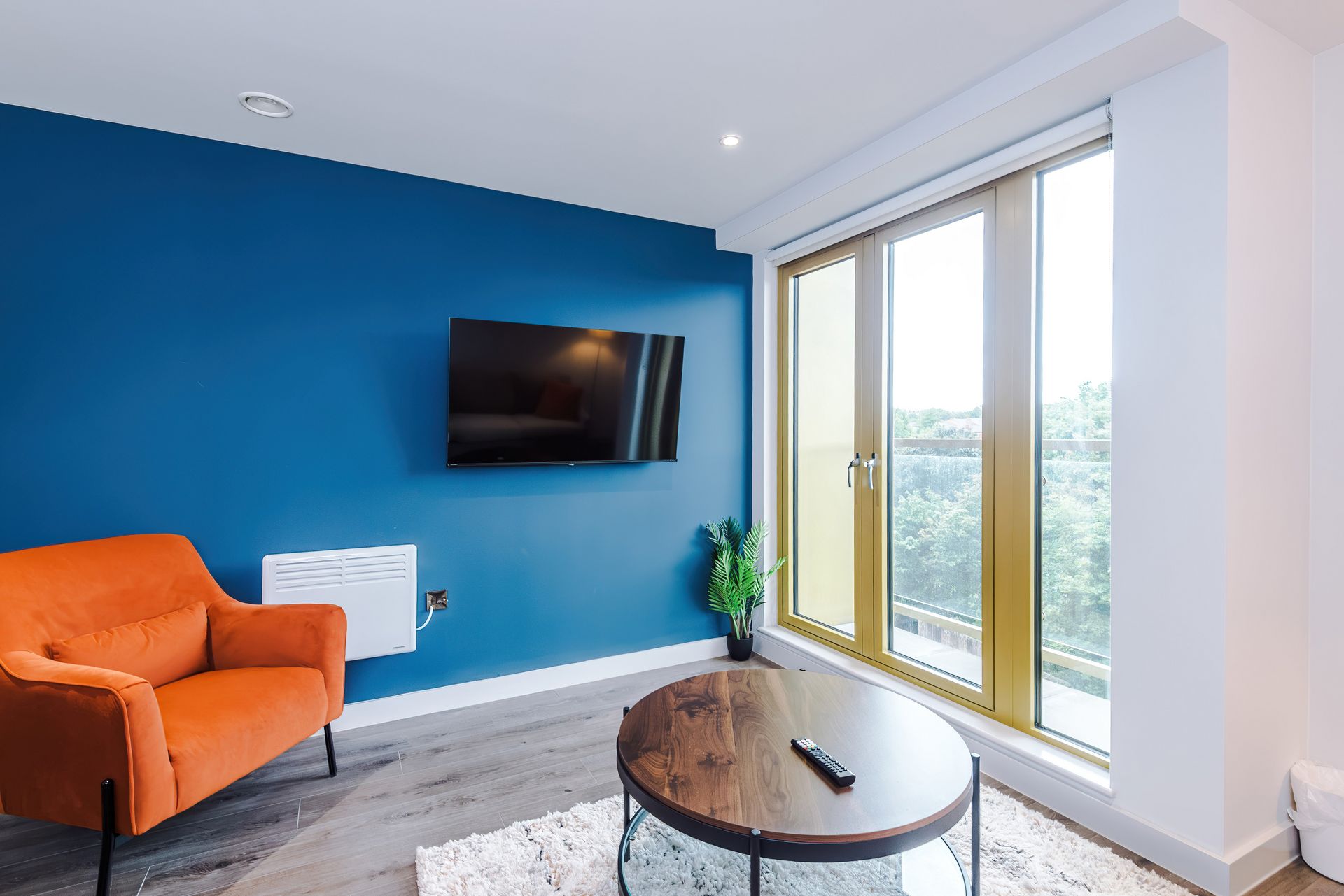 A modern living room with a blue wall, a flat-screen TV, a round wooden coffee table, and a large window with a view.