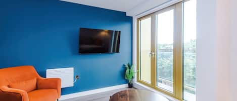 Ein modernes Wohnzimmer mit blauer Wand, einem Flachbildfernseher, einem runden Couchtisch aus Holz und einem großen Fenster mit Ausblick.