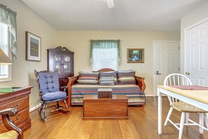 Una sala de estar acogedora, con piso de madera, un sillón mecedora, un sofá con ropa de cama a rayas, una mesa de centro de madera y una mesa de comedor con una silla.