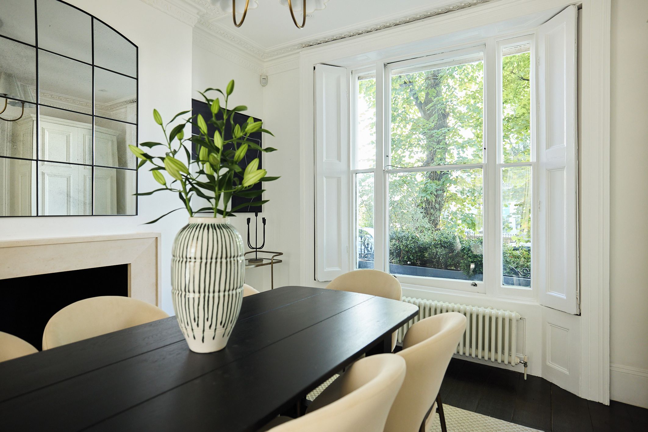 A dining area with a black table, white chairs, a vase with green leaves, a mirror, and a window with a view of trees.