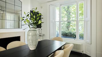 A dining area with a black table, white chairs, a vase with green leaves, a mirror, and a window with a view of trees.