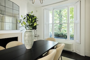 Ein Essbereich mit einem schwarzen Tisch, weißen Stühlen, einer Vase mit grünen Blättern, einem Spiegel und einem Fenster mit Blick auf Bäume.