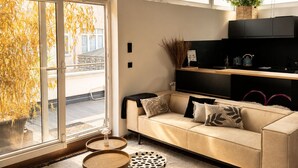 A modern living room with a sofa, a coffee table, and a side table. There is a large window with a view of trees and a building outside. - Stylish Bockenheim Apartment Near Messe, Restaurants & Vibrant City Life (Frankfurt am Main)