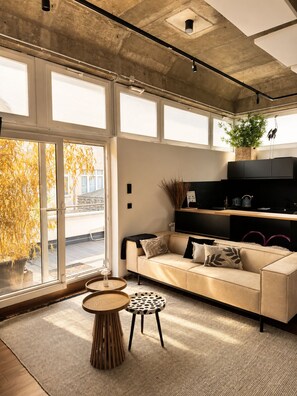 Ein modernes Wohnzimmer mit einer Couch, einem Couchtisch und einem Beistelltisch. Ein großes Fenster bietet Blick auf Bäume und ein Gebäude.