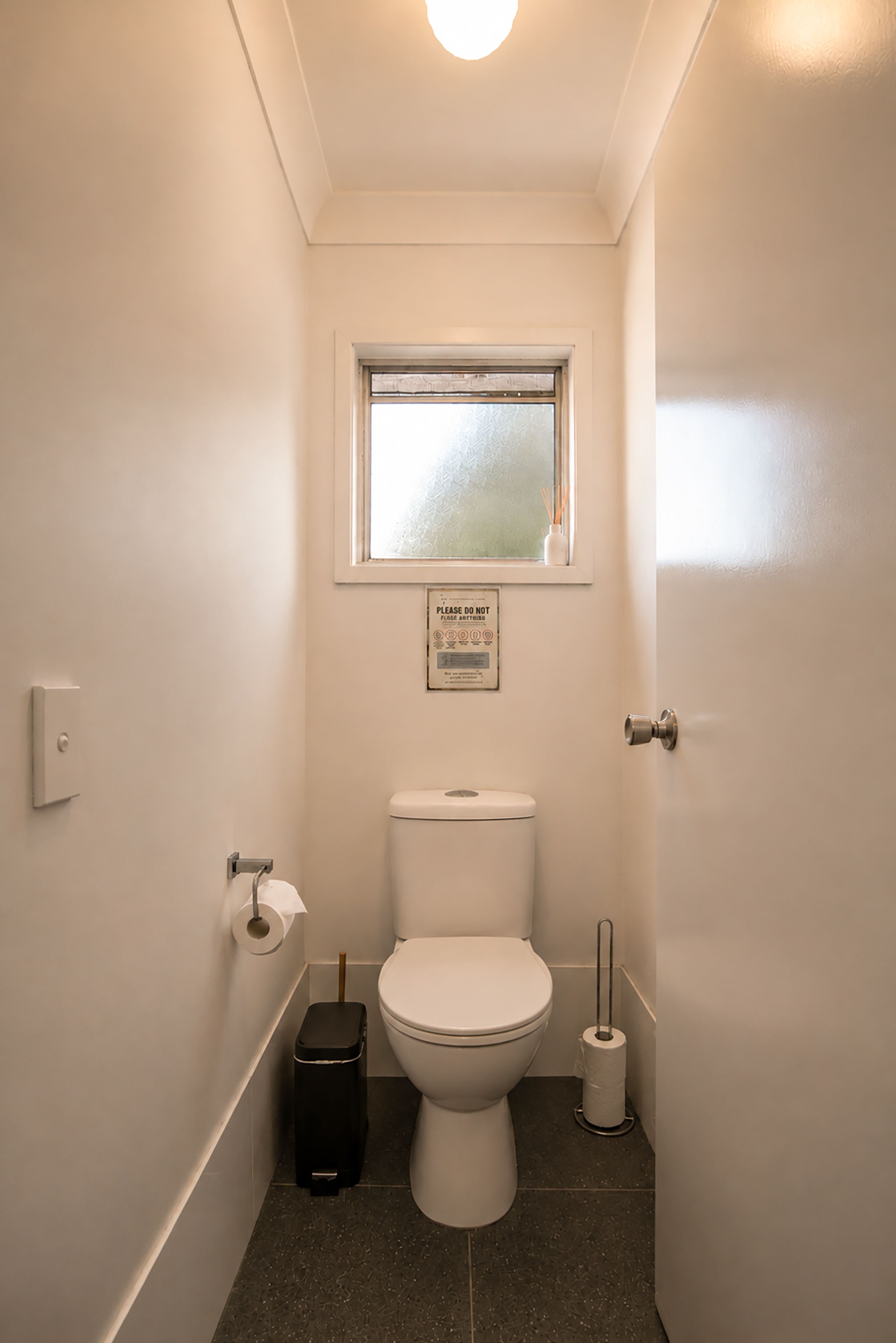 A small, narrow bathroom with a toilet, a window, and a trash bin.