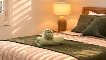 A neatly made bed with a green blanket and rolled towels, a bedside lamp, and a headboard with a woven texture.