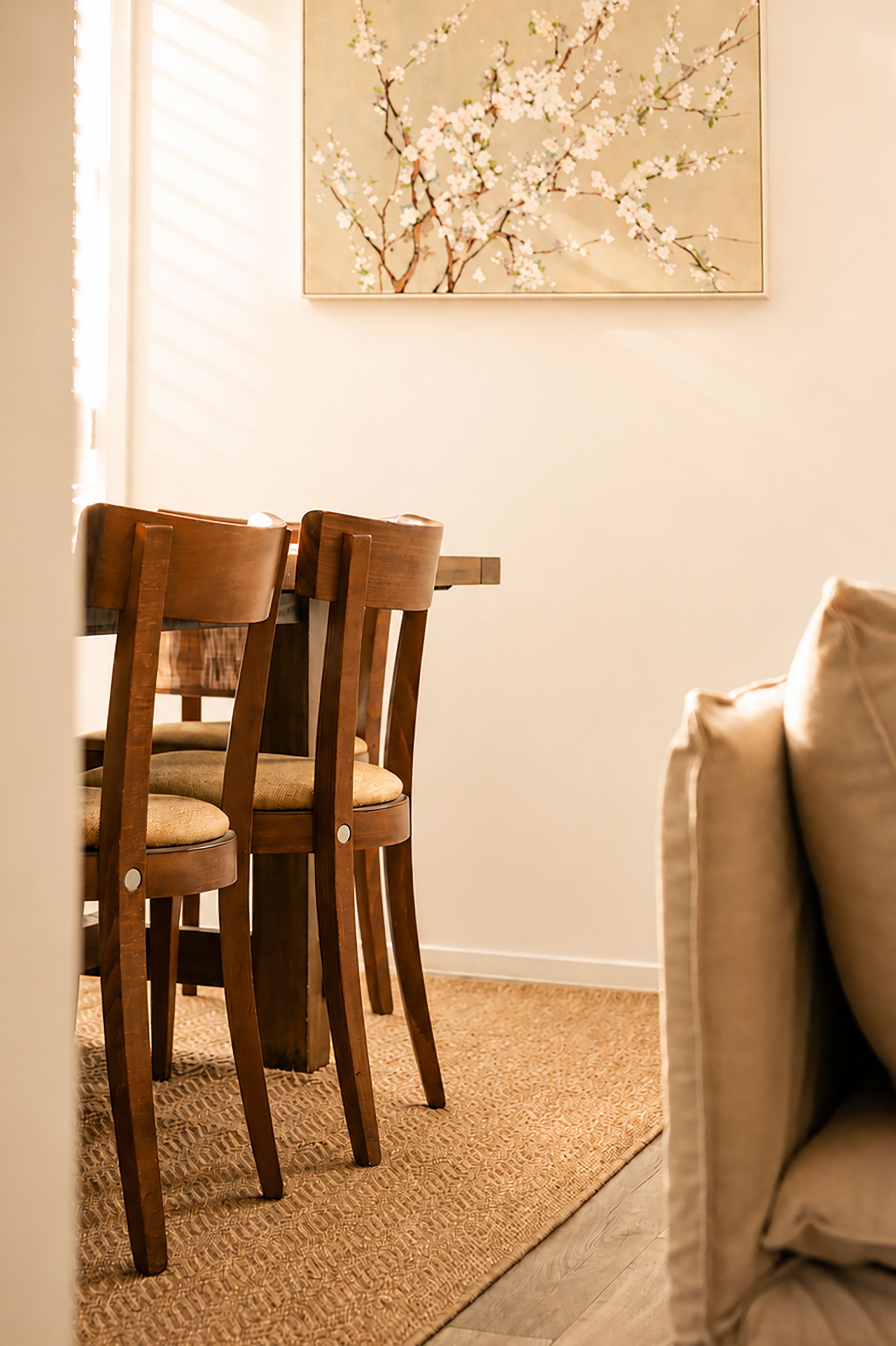 A dining area with wooden chairs and a table, a beige rug, and a wall-mounted painting.