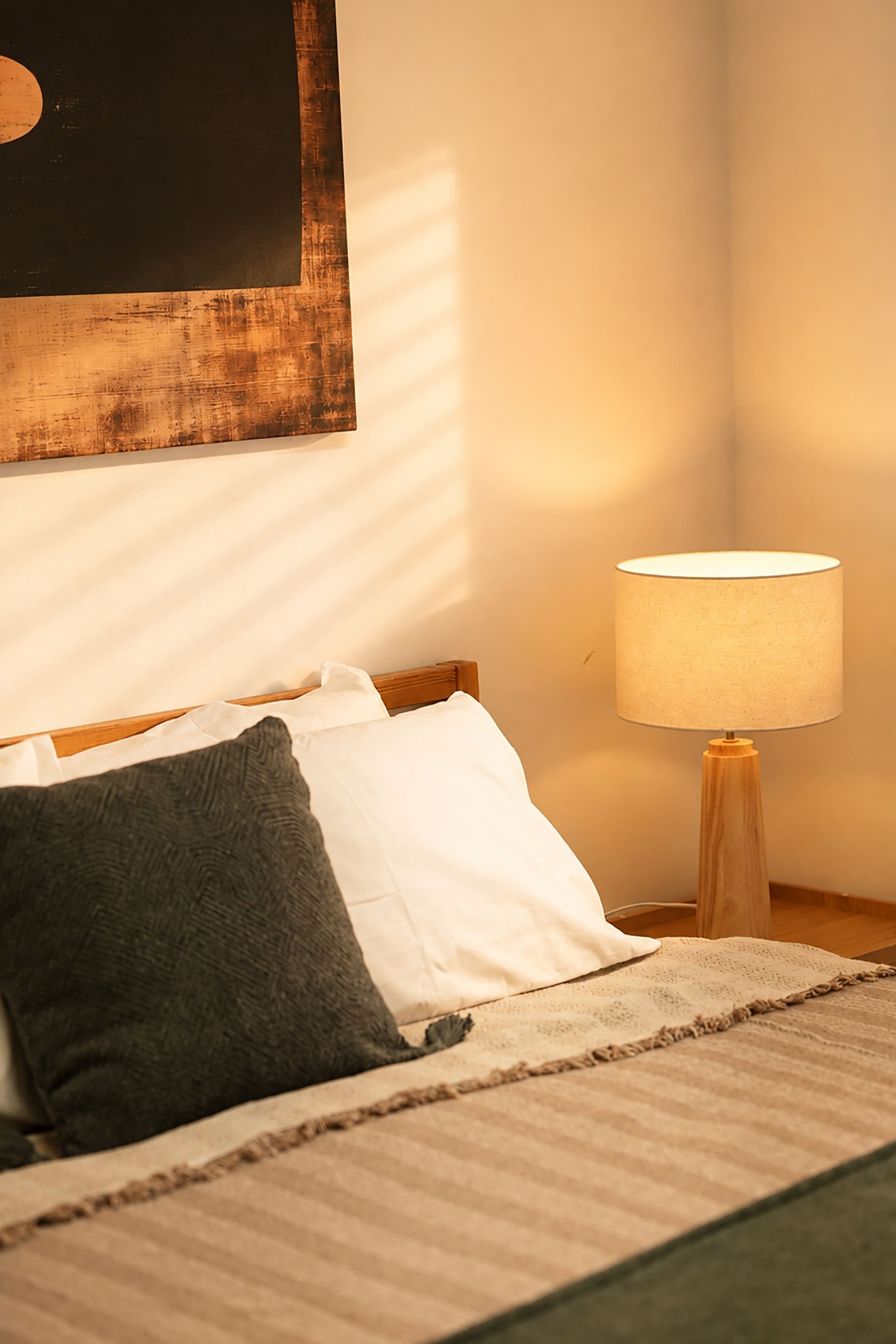 A neatly made bed with a white and gray blanket, a wooden nightstand with a lamp, and a wall with a framed artwork.