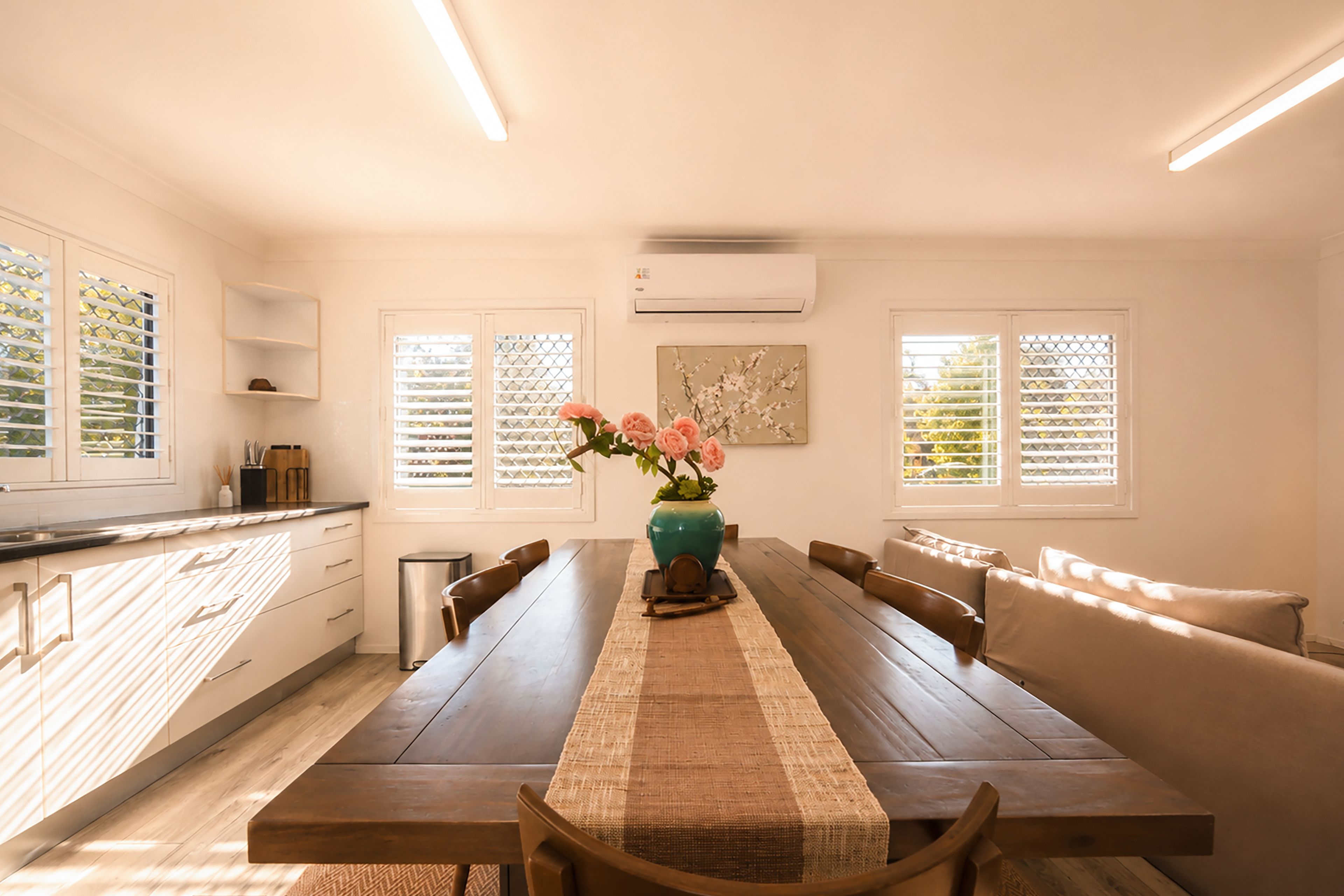 A modern kitchen with a dining area, featuring a long wooden table, chairs, and a vase with flowers.