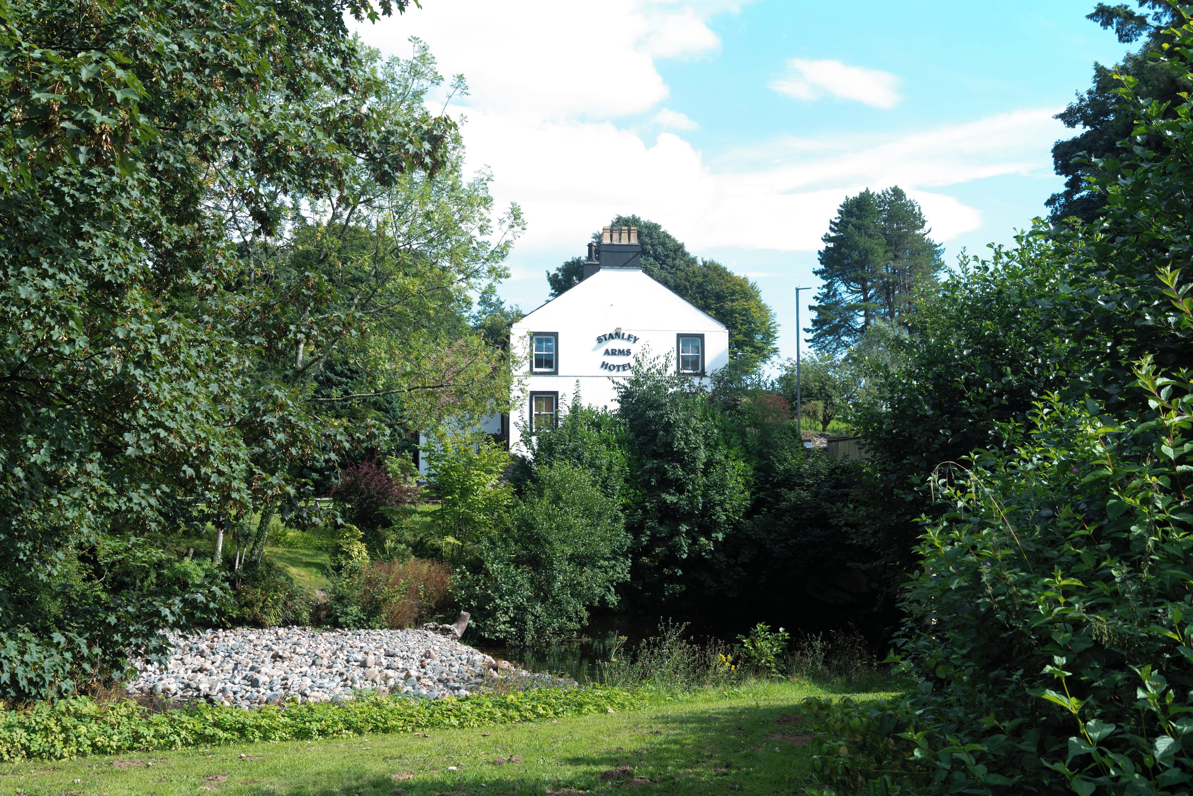 A white building with a sign reading 'Stanley Arms Hotel' surrounded by lush greenery and a stone pile.
