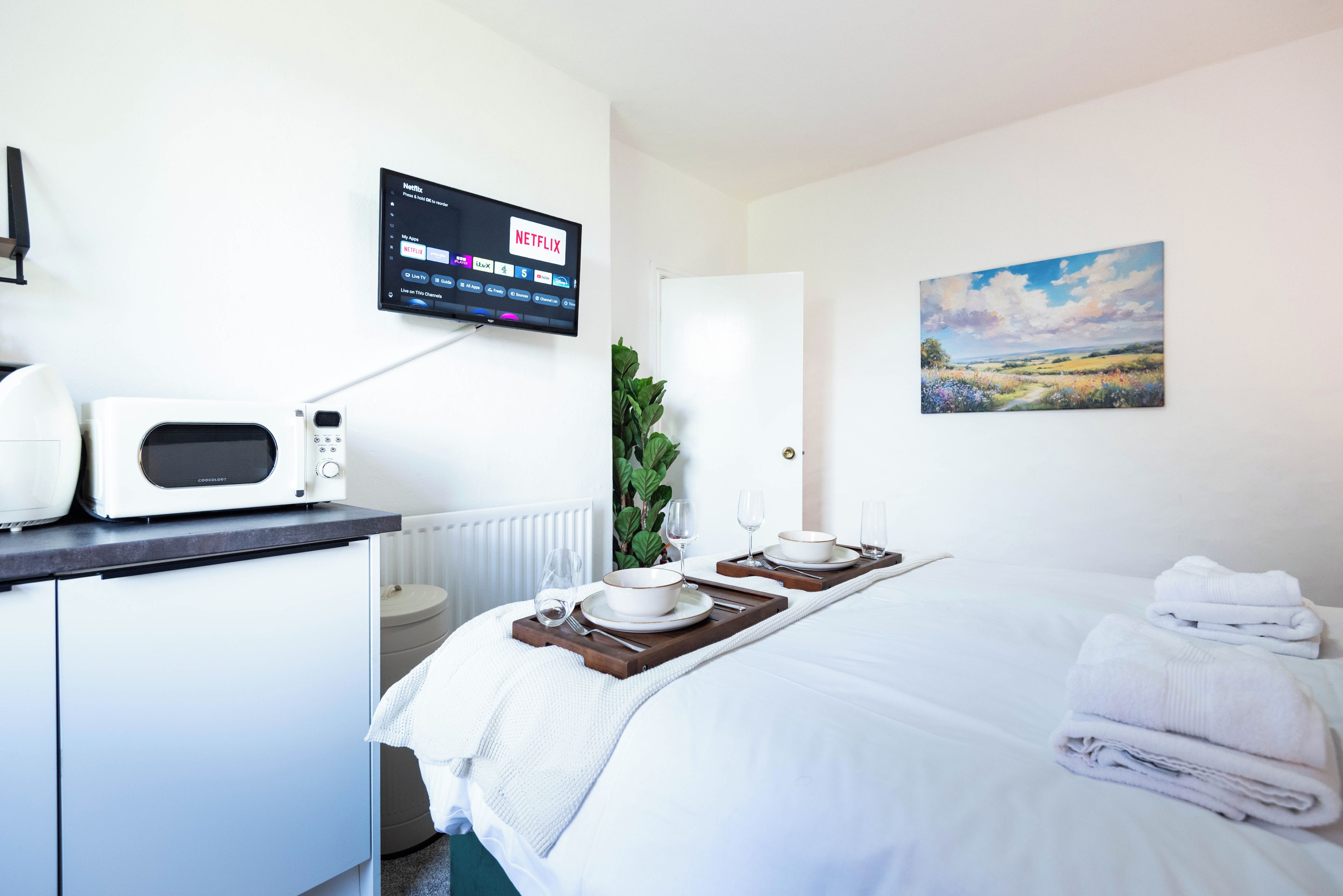 A hotel room with a bed, a bedside table with a microwave, a TV displaying Netflix, and a painting on the wall.