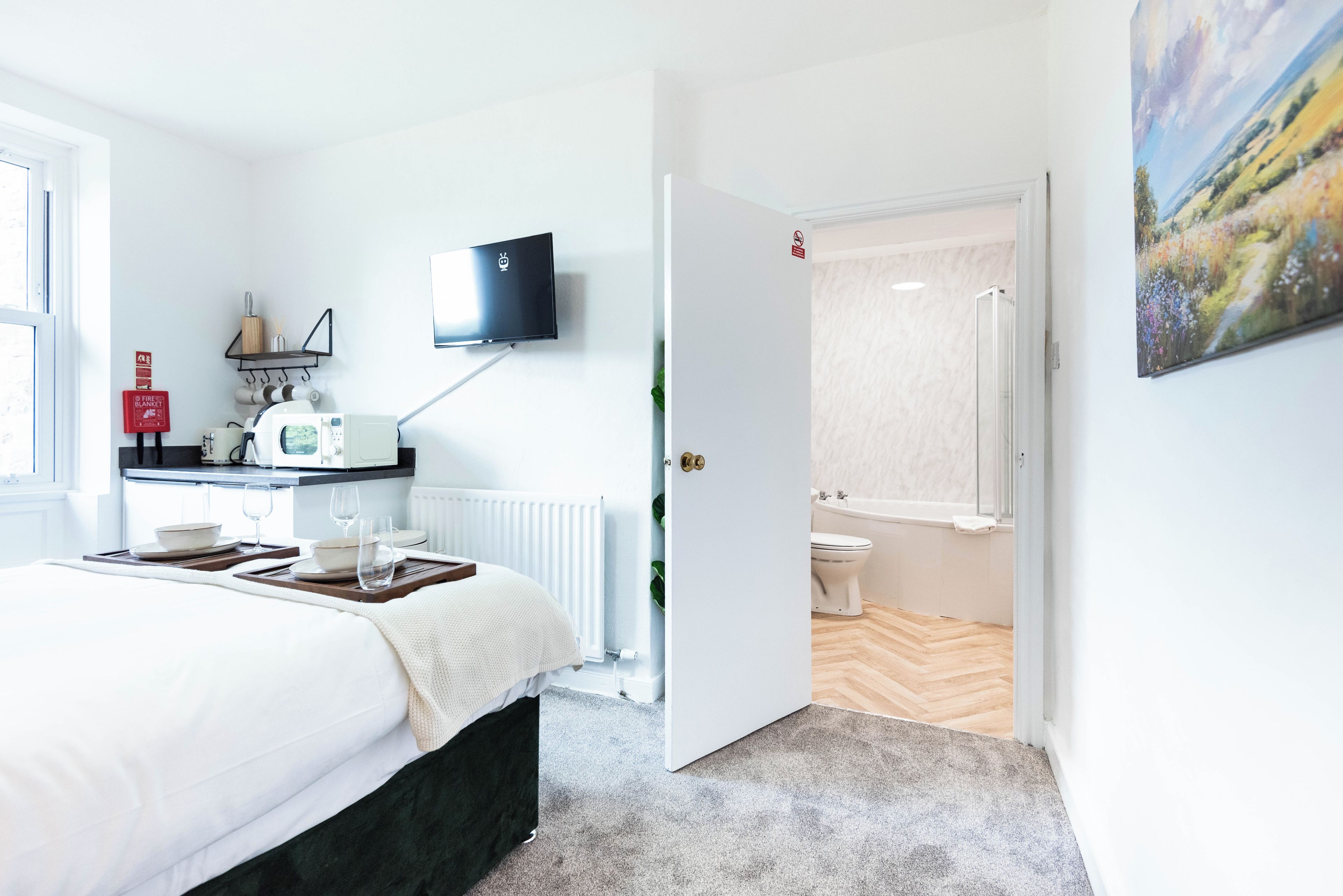 A hotel room with a bed, bedside table, a flat-screen TV mounted on the wall, a small kitchenette, and a bathroom with a toilet and shower.