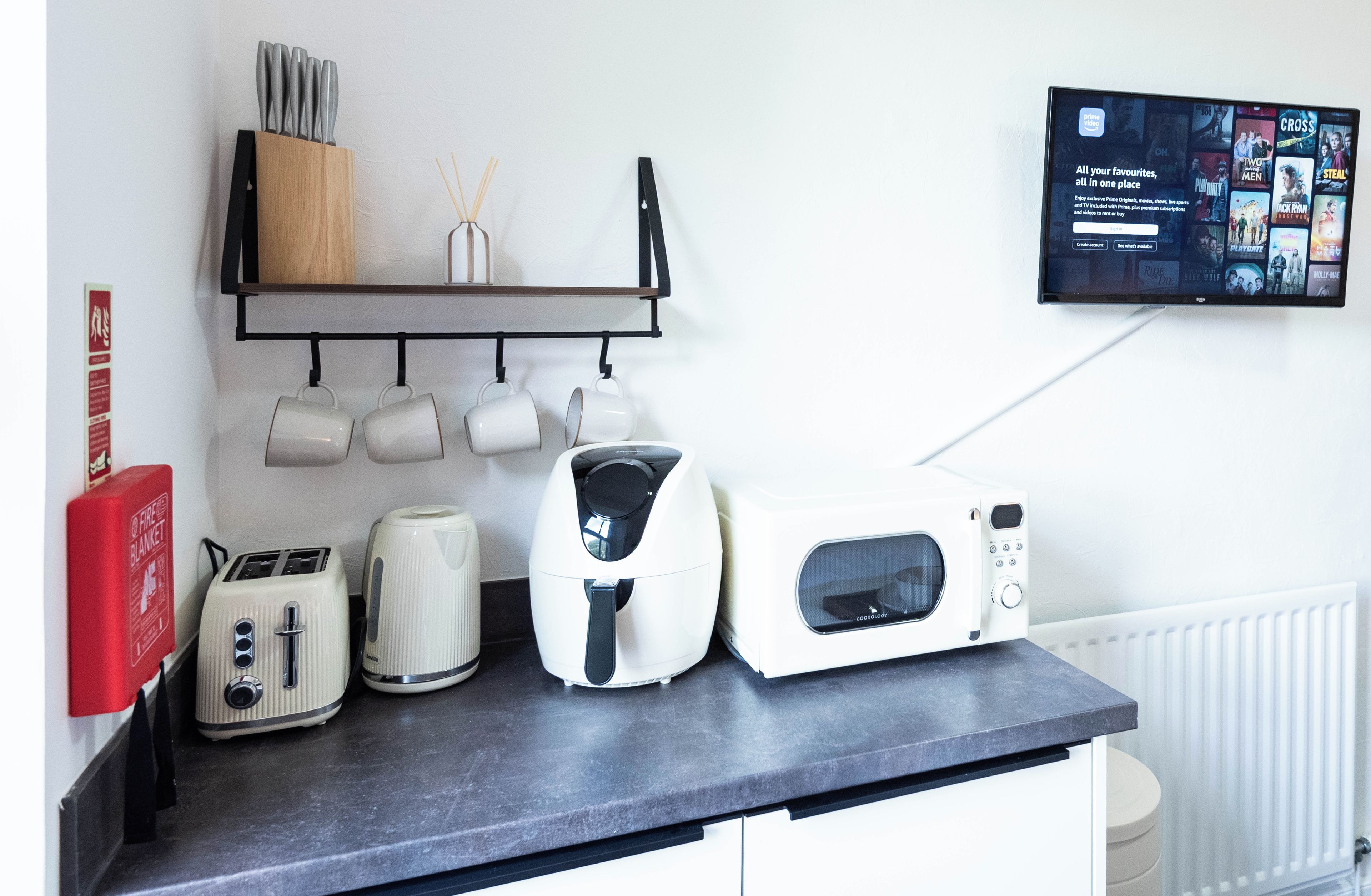 Una encimera de cocina con tostadora, hervidor y microondas, una manta anti-incendios y un televisor montado que muestra opciones de películas.