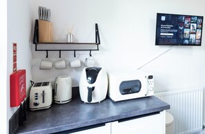 A kitchen counter with a toaster, kettle, and microwave, a fire blanket, and a mounted television displaying movie options.
