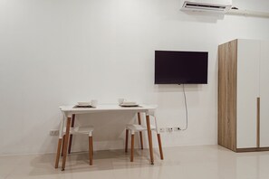 A minimalist room with a white desk, two chairs, a flat-screen TV mounted on the wall, and a wooden wardrobe.