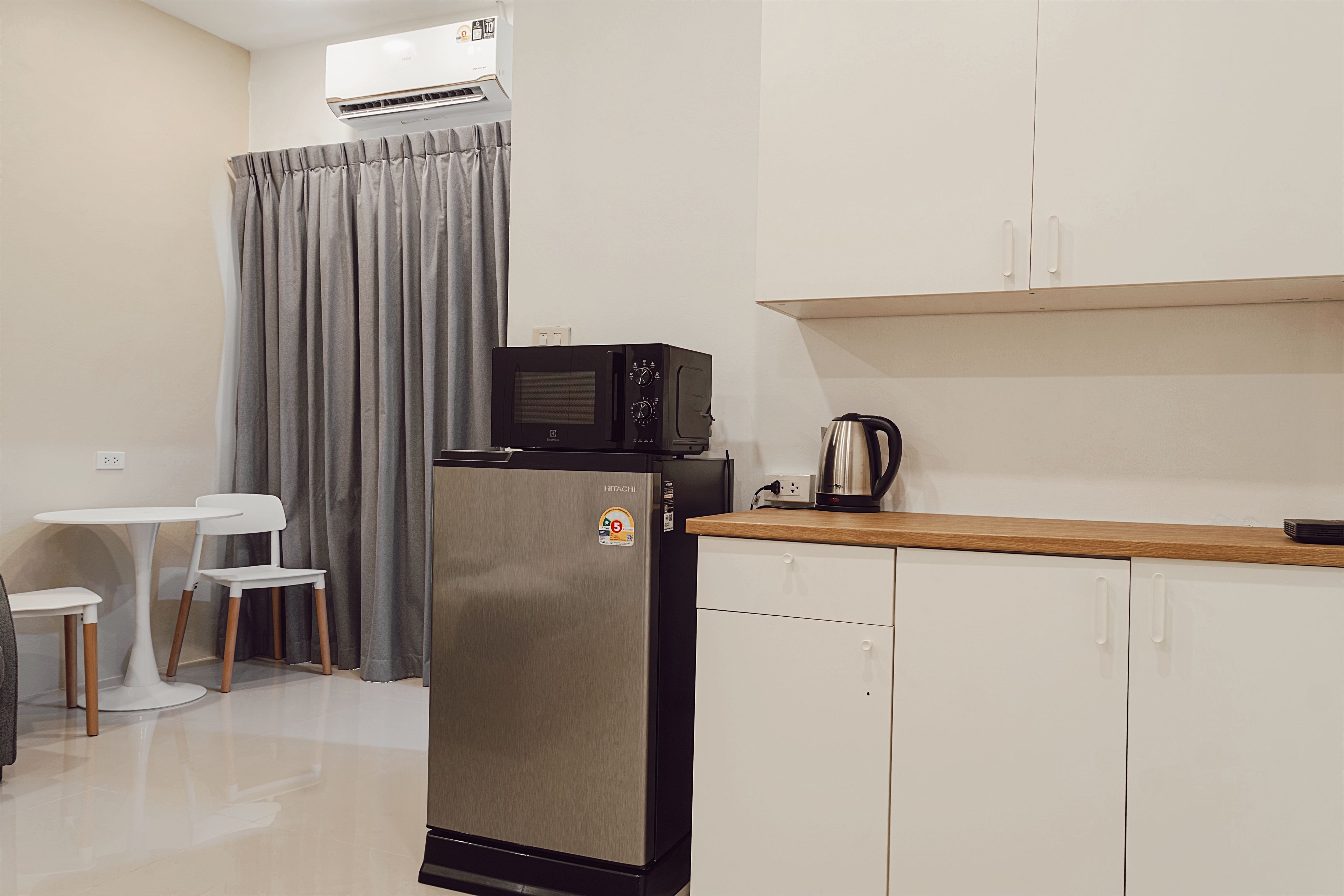 A compact kitchenette with a refrigerator, microwave, and kettle, a small dining table with chairs, and a grey curtain.
