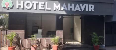 Entrada de un hotel con un letrero que dice 'HOTEL MAHAVIR', asientos al aire libre con plantas en macetas y una fachada de edificio moderna.