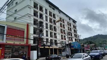 Un edificio de varios pisos con locales comerciales en la planta baja, un estacionamiento y un paisaje montañoso al fondo.