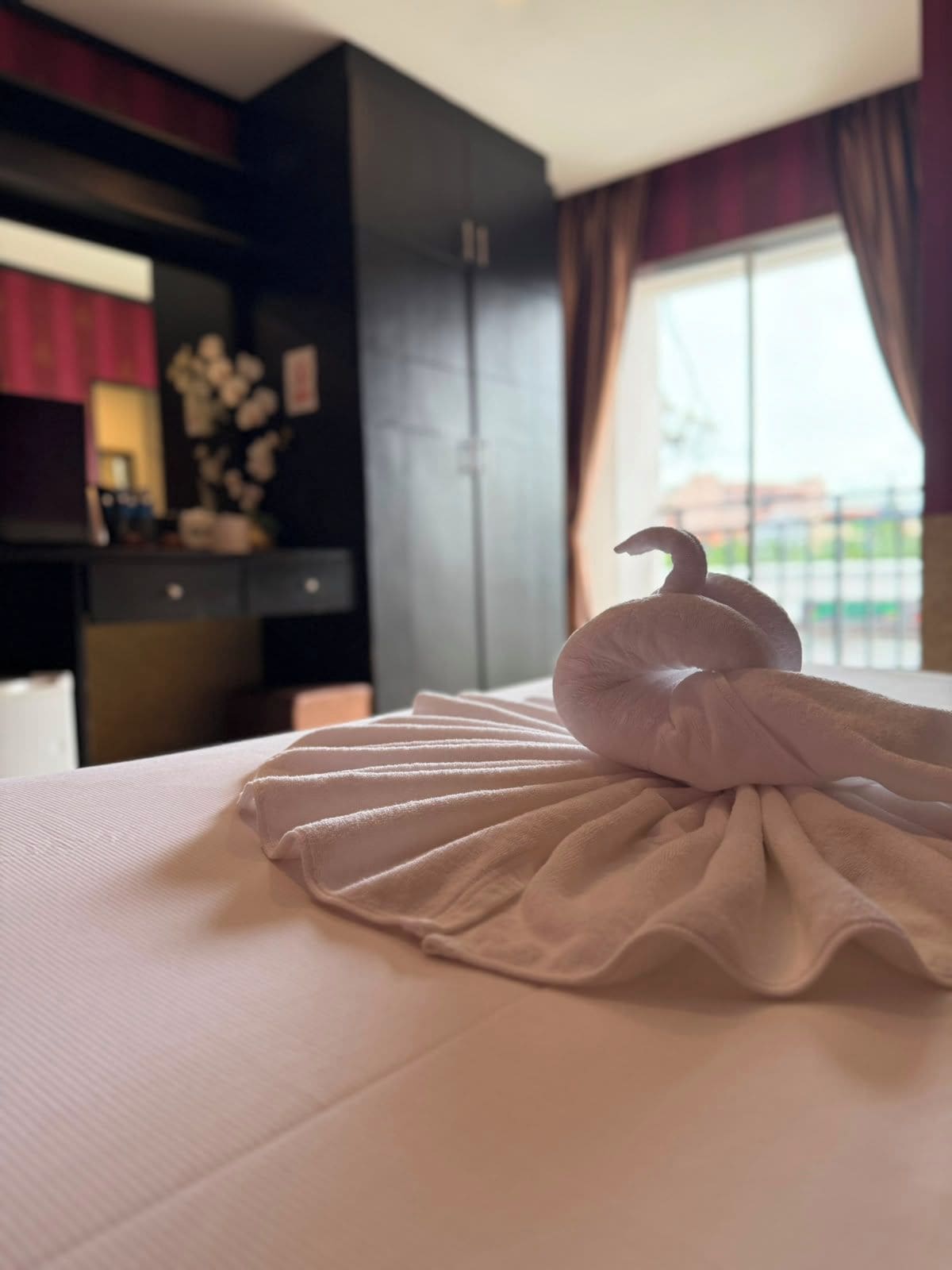 Una cama bien hecha con una toalla doblada en forma de cisne, un armario de color oscuro y una ventana con cortinas.