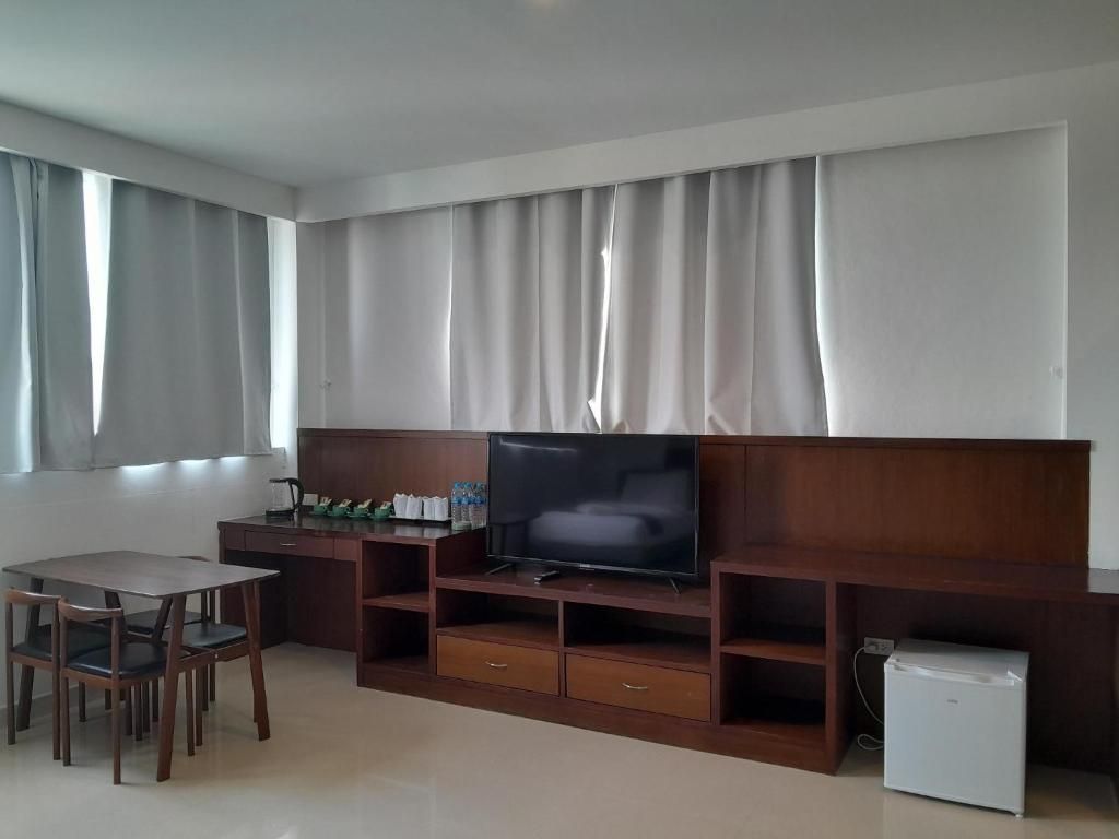 Una sala de estar con un televisor de pantalla plana sobre un soporte de madera, un refrigerador pequeño y un comedor con sillas.