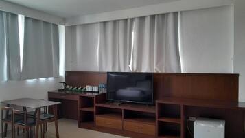 Una sala de estar con un televisor de pantalla plana sobre un soporte de madera, un refrigerador pequeño y un comedor con sillas.