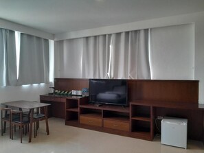 Una sala de estar con un televisor de pantalla plana sobre un soporte de madera, un refrigerador pequeño y un comedor con sillas.
