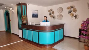 Un mostrador de recepción con una persona sentada detrás, un ventilador de pared y decoración en la pared.