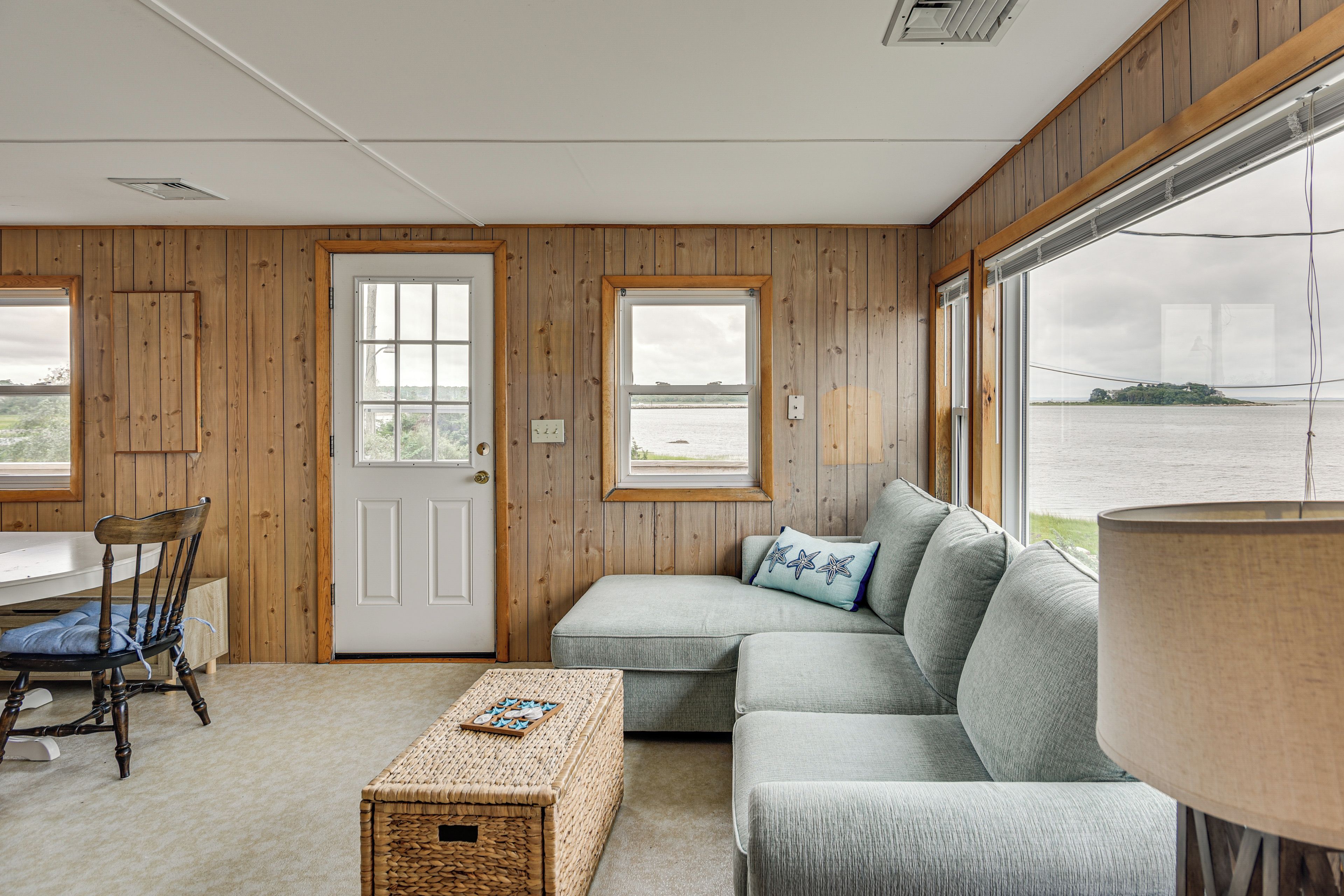 A room with wooden walls, a white door, a grey sofa, a wicker coffee table, and a view of a body of water through large windows.