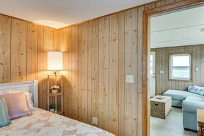 Una habitación con paneles de madera, una cama con ropa de cama floral, una lámpara de noche, una planta pequeña y vistas a la costa a través de una ventana.
