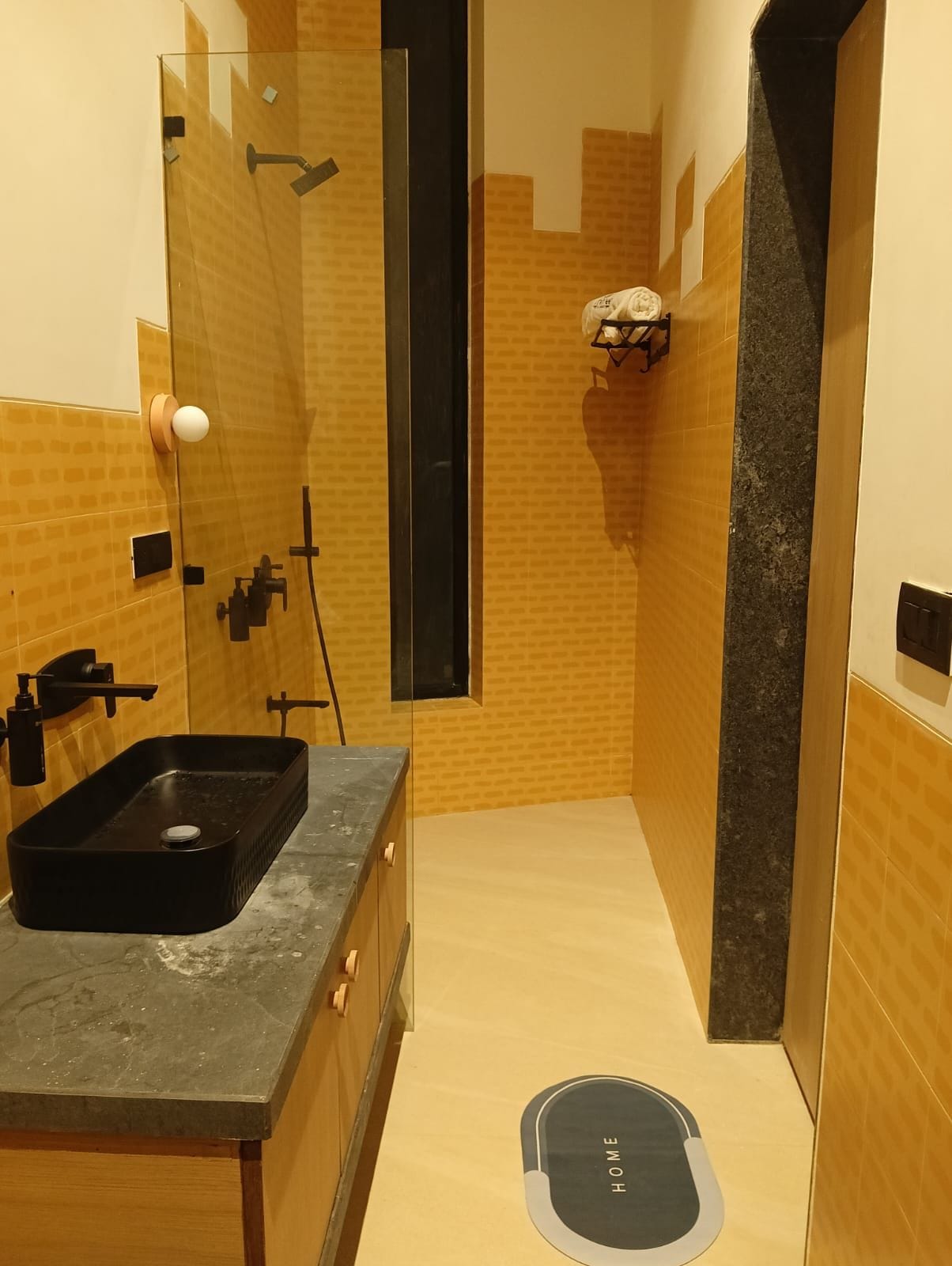 A modern bathroom with a glass shower enclosure, a black sink, and a 'HOME' mat on the floor.