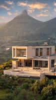 A modern two-story house with a flat roof and large windows, surrounded by lush greenery and a mountain in the background.