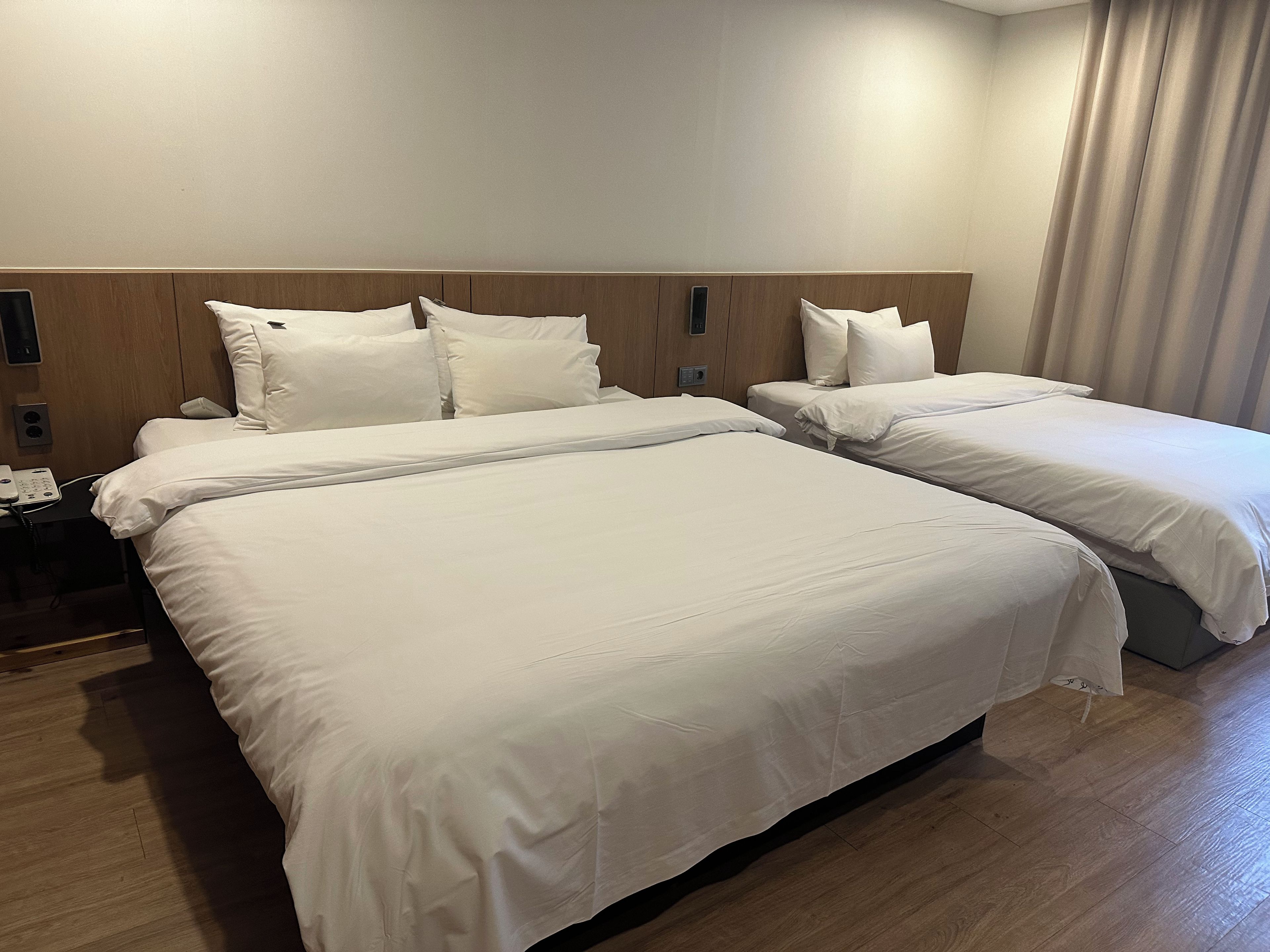 Quarto de hotel com duas camas, cabeceira de madeira, mesa de cabeceira com telefone e cortina.