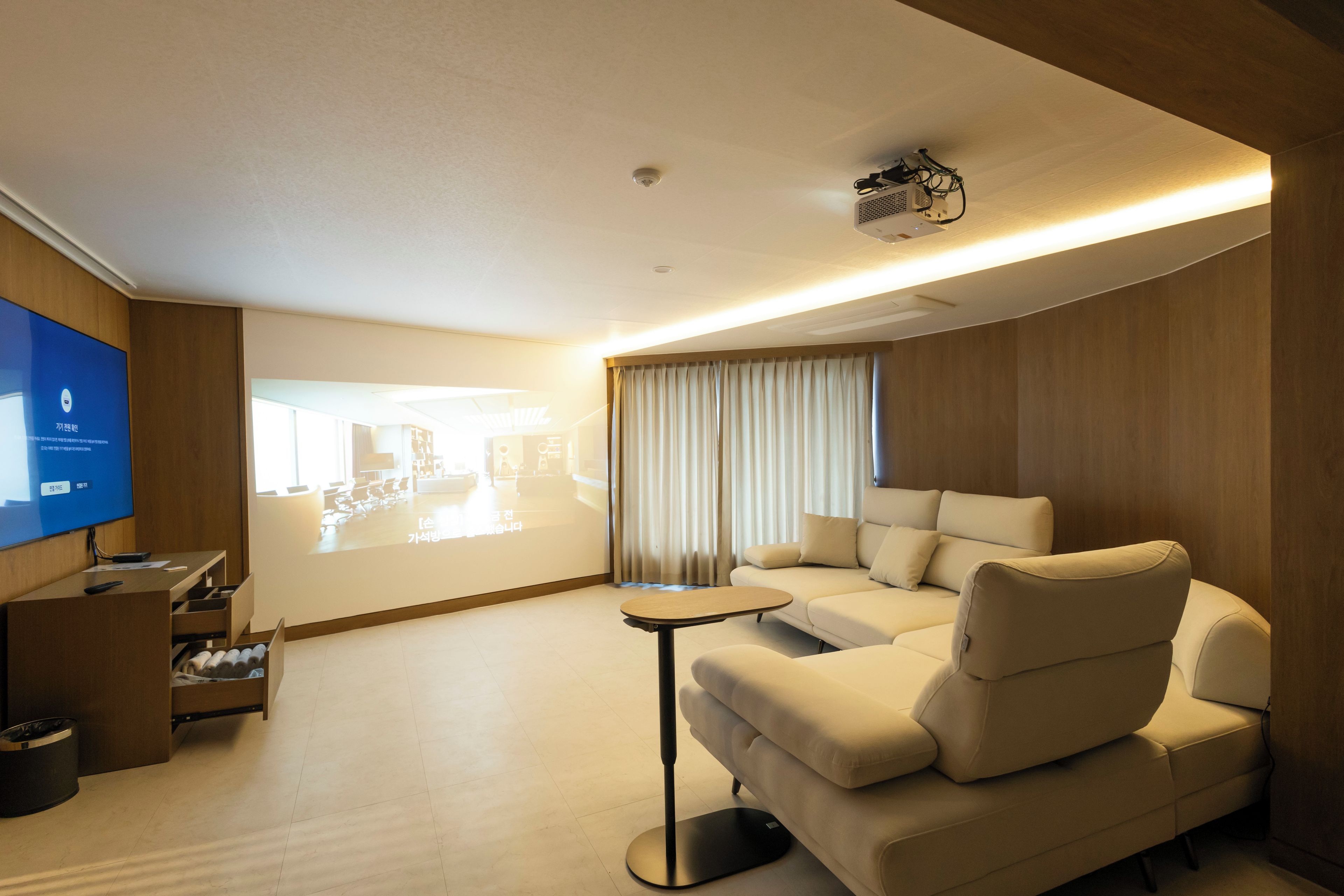 Een moderne hotelkamer met een grote flatscreen-tv, een bank, een klein tafeltje en een projectorscherm waarop een plattegrond staat.