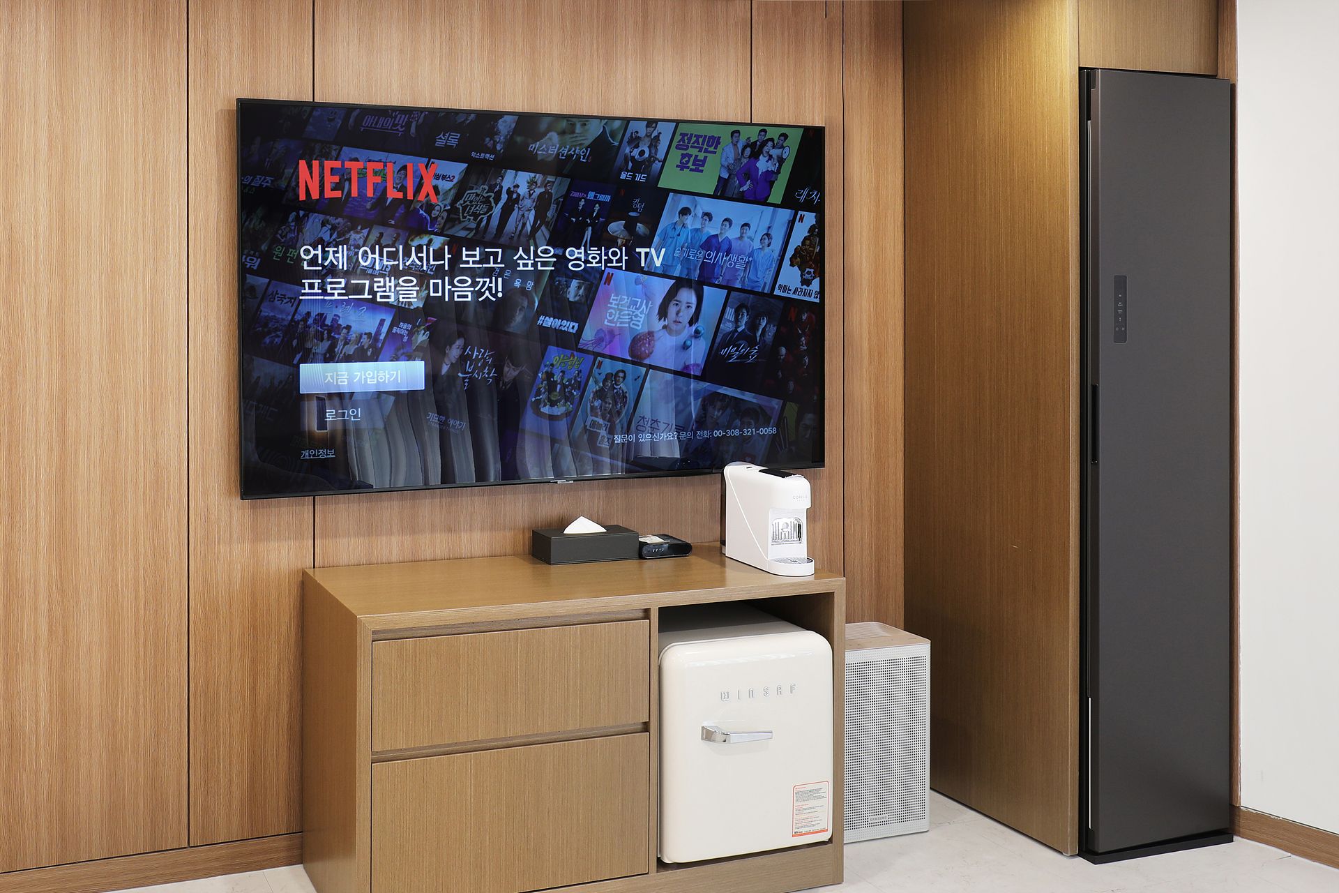 Una sala de estar moderna con un televisor de pantalla plana montado en una pared de madera, un refrigerador blanco pequeño y un mueble negro.