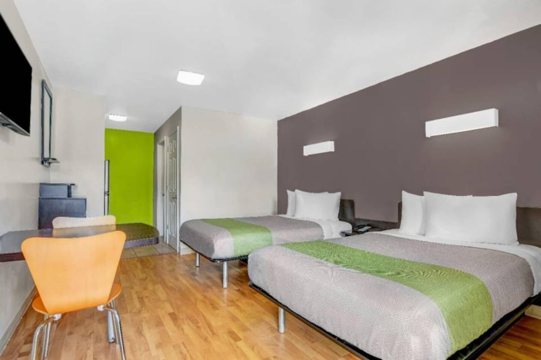 A hotel room with a wooden floor, a single orange chair, two beds with grey and green bedding, a grey headboard, and a television mounted on the wall.