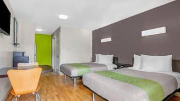 A hotel room with a wooden floor, a single orange chair, two beds with grey and green bedding, a grey headboard, and a television mounted on the wall.