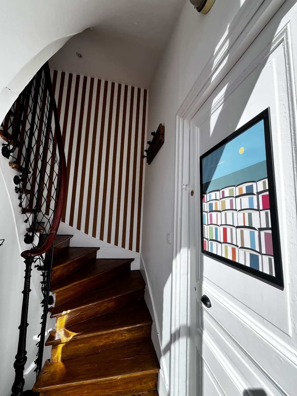 Un escalier en bois avec une rampe noire menant à une porte blanche ornée d’une fenêtre aux couleurs vives.
