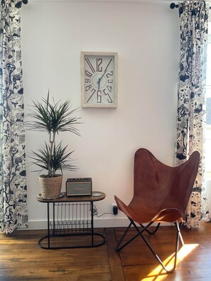 Ein Zimmer mit Holzboden, einem braunen Sessel, einem Topfpflanzen, einem kleinen Tisch mit einem Radio und einer Wanduhr.