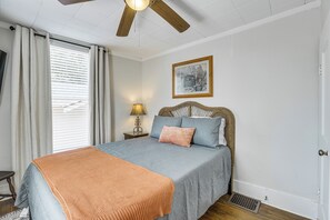 Een slaapkamer met een bed, nachtkastje, lamp, raam met gordijnen en een plafondventilator.