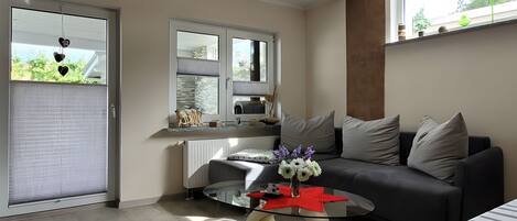 A modern living room with a glass coffee table, a gray sofa, and a large window with blinds.