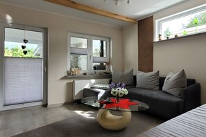 A modern living room with a glass coffee table, a gray sofa, and a large window with blinds.